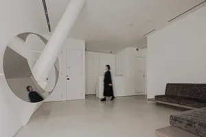 Interior of a modern, minimalist living room with concrete floor and walls, featuring a dark-colored sofa and a person walkin