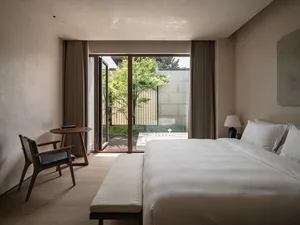 A minimalist bedroom with a white fabric bed on a wooden floor, featuring natural light and greenery outside through large wi