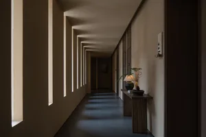 A minimalist corridor with concrete walls and floor, featuring rectangular windows and a wooden side table with a lamp and po