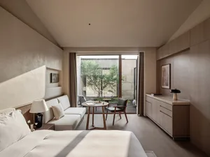 A minimalist bedroom featuring wooden furniture and textured textiles, with a view of greenery through large windows.