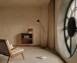 A modern living room with minimalist design, featuring a wooden armchair, a floating wooden shelf, a standing lamp, and a rou