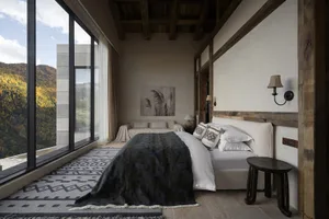 A modern minimalist bedroom with white-on-white bedding, a wooden headboard, and a large window overlooking the mountains.