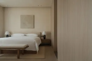 A minimalist bedroom with a bed, nightstands, and a bench, all in neutral colors and featuring a wooden headboard and a large