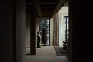 A person dressed in traditional attire standing in a minimalist architectural interior with exposed wooden beams and large wi