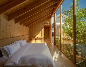 A minimalist bedroom with wooden walls and floor, featuring a comfortable bed with white bedding and a large window overlooki