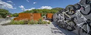 Bicycles parked on a gravel surface with a metal fence and greenery in the background.