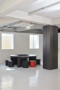 A modern industrial conference room featuring a black circular table, red stool, and concrete space.