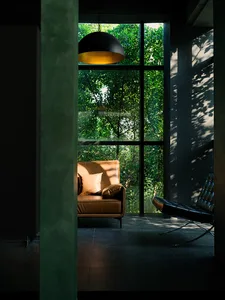 Modern minimalist living room featuring a brown leather sofa with a hanging light fixture, large windows showing lush greener