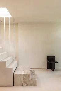 A minimalist interior space with concrete steps, a marble bench, and a black chair against a plain wall.