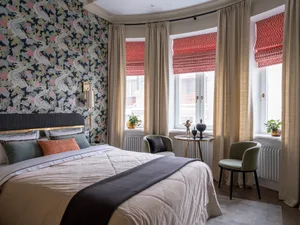 A modern bedroom with a patterned wallpaper wall and large windows with red patterned blinds.