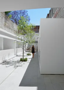 A minimalist, white interior with concrete walls and floors, featuring a single tree and a woman walking.