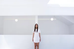 Woman standing in a minimalist bedroom with neutral colors and white walls.