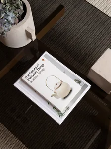 A white book titled 'The Beauty of Everyday Things' featuring a teapot illustration, placed on a glass coffee table with a po