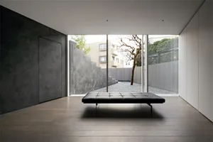 A modern minimalist bedroom with a concrete floor, black upholstered seating area, and a view of a tree-lined yard.