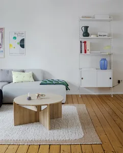 A modern minimalist living room featuring a light-colored oak wood coffee table, a grey couch, and a white wire shelving unit