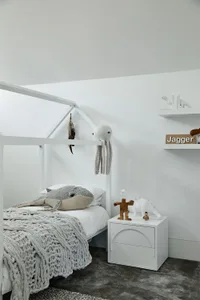 A minimalist kids bedroom featuring a white bed with a grey blanket, a white nightstand with dinosaur toys, and a white frame