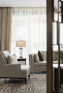 A modern neutral-toned living room with a light-colored armchair and a sofa adorned with patterned cushions, featuring a side