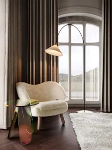 A cozy living room with a white fur chair, a modern glass side table, and a floor lamp with a view of mountains through large