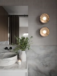 Modern bathroom interior featuring marble tiles, a round marble sink, and two round wall lights with a vase of greenery and a