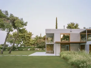 Modern house with large windows, glass walls, and a concrete patio surrounded by greenery.