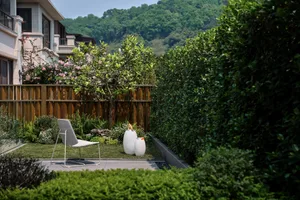 A modern outdoor space featuring a patio with a white chair, white vases, and lush greenery.