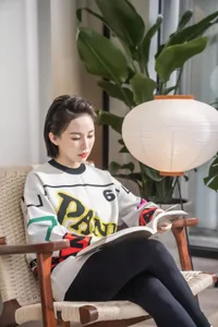 A woman wearing a colorful sweater and black leggings reads a book on a woven wicker armchair near a potted plant and floor l
