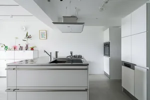 A modern, minimalist white kitchen with a cooking area in the center, featuring a double-sided stainless steel countertop, an