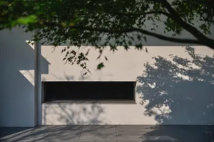 Modern exterior of a building with white walls and concrete materials, framed by tree shadows.