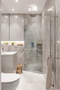A minimalist bathroom featuring a grey marble shower enclosure with glass doors, a white porcelain toilet, and a sleek white 