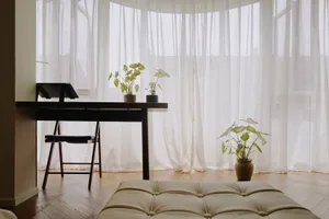 A cozy home office featuring a desk, chair, and potted plants under white curtains.