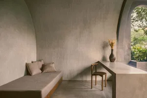Modern minimalist living room with textured concrete walls and furniture, featuring a simple wooden chair with a vase of drie