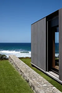 Modern seaside residence featuring metal cladding and concrete, with a large glass window overlooking the ocean.
