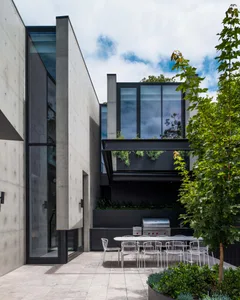 A modern residential patio featuring concrete flooring, glass walls, and a dining area with greenery and outdoor kitchen.