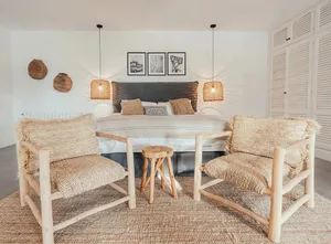 Modern minimalist bedroom featuring a bed with dark headboard, two wooden armchairs with woven rattan cushions, and a wooden