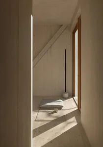 Minimalist bedroom with wooden flooring and walls, a white bed, and sunlight streaming through the window.