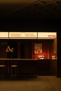 A casual bar with a brick wall, two chairs, and a counter with wine glasses, a cup, and a bowl, with a man standing behind the counter