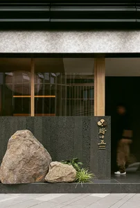 A modern restaurant with a traditional Asian design, featuring a black facade with a rock wall and a wooden door, with a dining table and chair in the center, and two people standing on either side