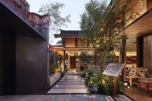 Traditional Asian-style house with a wooden structure, tiled roof, and stone walkway, surrounded by potted plants and featuring arches and balconies