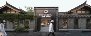 A man walks in front of an Asian restaurant with a black facade, featuring dining tables and chairs inside, creating an inviting and comfortable atmosphere