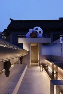 A serene Asian-style building with a stone panda statue on a balcony, overlooking a bridge and street below, surrounded by benches in a peaceful environment