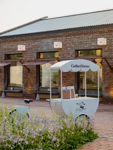 A brick building with an outdoor seating area featuring two umbrellas, a bicycle, and a motorcycle, with two benches underneath the second umbrella
