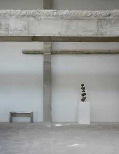 Empty industrial space with concrete walls and floors, featuring three sculptures and a bench, with a minimalistic and stark atmosphere