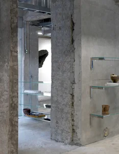 A well-lit art gallery with a display case filled with vases, bowls, and books, set against industrial concrete walls and illuminated by overhead lights