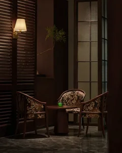 A darkly lit living room with two chairs and a small table, surrounded by potted plants and a bottle, creating a cozy atmosphere