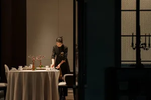 A woman in an apron prepares food on a table in a professional kitchen with bowls, a spoon, and a knife