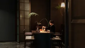 A woman sits at a dining table in a darkly lit room surrounded by chairs, creating a cozy and intimate atmosphere