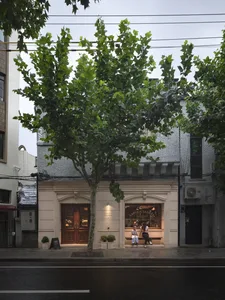 A serene outdoor scene of a building with a tree in front, featuring a white facade, two people, and a bench, set against a calm atmosphere