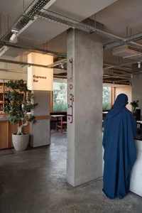 A modern cafe interior with concrete walls, wood accents, and metal elements, featuring a woman standing at a counter and several chairs for seating, with a potted plant in the background