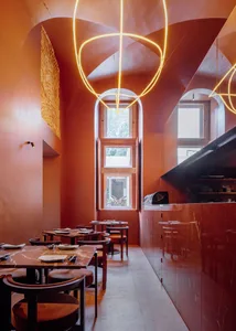 A dimly lit modern restaurant with red walls, clean lines, and minimal decoration, featuring several tables and chairs arranged throughout the space