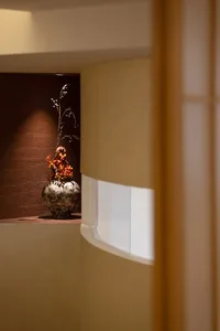 A living area with a vase of yellow flowers on a table in front of a window, surrounded by a potted plant and natural light